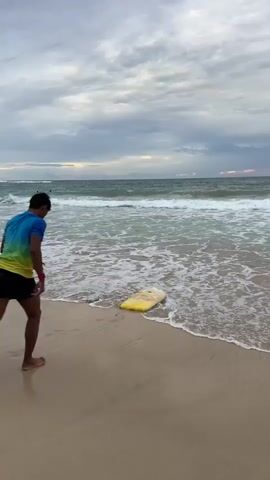 A person on a beach walks towards a yellow boogie board partially submerged in the shallow water at the edge of the ocean. The person is wearing a blue and green gradient t-shirt and dark shorts. They step onto the boogie board, which causes it to lift and glide slightly on the water. The person then stands up on the boogie board and rides it a short distance before falling off. The ocean has waves rolling in under a cloudy sky.