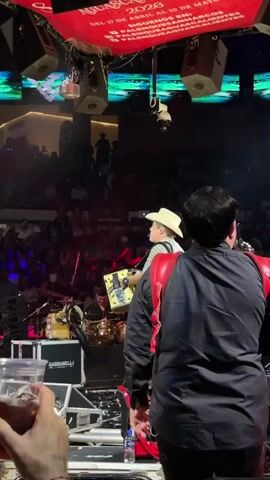 The image depicts a live music concert taking place in an indoor arena at night. On stage, a band is performing. The central figure is a man in a cowboy hat, playing a gold Gabbanelli accordion, with his back mostly to the viewer as he looks up. Another band member wearing a black shirt and red harness is partially visible to the right. A drummer is seated behind a drum kit on the left side of the stage. Microphones, speakers, and stage lights are arranged around the performers.

A large audience fills the tiered seating in the background, dimly lit with some visible cell phone lights.

Overhead, a prominent red banner displays text in white: "AGUASCALIENTES," "MARCOS," "2023," "DEL 17 DE ABRIL AL 10 DE MAYO," "PALENQUE," "FERIA DE SAN MARCOS," and "AGUASCALIENTES." This indicates the event is part of the San Marcos Fair in Aguascalientes, held from April 17th to May 10th, 2023, likely at a palenque venue. A security camera is also visible hanging from the ceiling structure.

In the immediate foreground, a person's hand holds a clear cup containing a dark beverage. The scene is illuminated by the stage lighting, highlighting the performers and equipment.