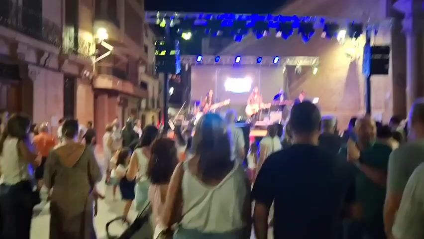 The video shows an outdoor night concert with a live band performing on a stage. The stage is set up in a street between buildings, with lighting and speakers visible. A crowd of people are gathered in front of the stage, watching the performance. The buildings on either side of the street are lit up, and there are some architectural details visible, such as balconies and windows. The atmosphere appears to be festive and lively.
