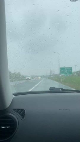 The view from inside a car driving on a wet road, with raindrops visible on the windshield. The windshield wipers are active, clearing the view. Streetlights line the road, and a green road sign is visible on the right. Other cars are driving in the distance. The car's dashboard is visible in the foreground. Music is playing.