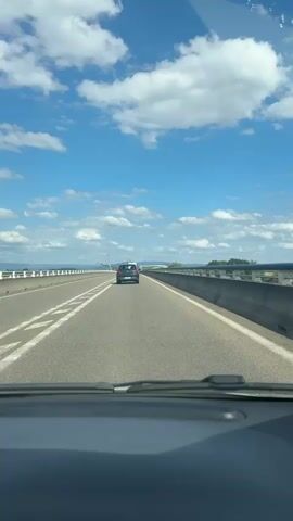 A car is driving on a highway. The camera is inside another car, looking out the windshield. The sky is blue with white clouds. There is a highway with lanes marked by white lines. A concrete barrier runs along the right side of the road. Ahead, another car is visible, driving in the same direction. Further ahead, there is a blue sign with stars, and a speed limit sign of 70. There are trees and hills in the distance. The car in front appears to be black. The license plate of the car in front is visible and appears to read "BC 355 SX". There is a white object on the roof of the car in front.