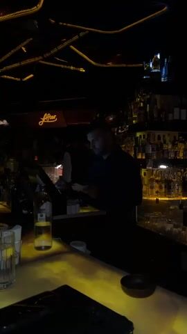 A man is standing behind a bar in a dimly lit establishment. He is wearing a dark shirt. The bar is stocked with numerous bottles of alcohol on shelves behind him. In the foreground, a bottle of liquor, a glass, and an ashtray are visible on the counter. A digital menu or ordering system is illuminated on the bar in front of the man. The lighting is predominantly yellow and orange, creating a moody atmosphere. The word "After" is visible in neon letters above the bar.