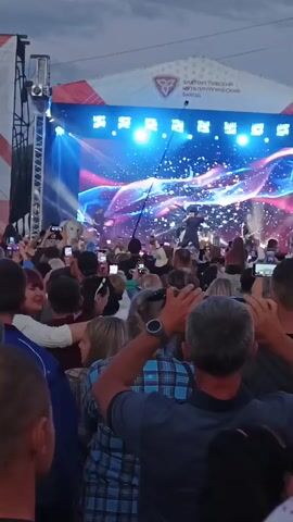 The video shows a live music performance on an outdoor stage. A crowd of people are visible in the foreground, holding up their mobile phones to record the event. The stage is lit with bright blue and pink lights, and there are pyrotechnic displays occurring. A band is performing on stage, with a lead singer in the center. The background features a large banner with text and a logo. The overall atmosphere appears to be energetic and celebratory.