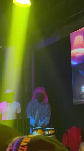 Here's a detailed description of the video:

The video shows a dimly lit nightclub with a performance stage where two musicians are performing. The first musician, wearing a white shirt and white cap, is likely DJing or managing sound equipment. The second musician is actively playing a set of bongos with drumsticks while wearing a hooded sweatshirt and a bright pink furry hat. 

The stage lighting changes frequently, with spotlights in various colors like yellow, red, and purple flashing and moving across the stage and the audience. The audience is visible in the foreground, with people wearing different colored shirts and glasses, suggesting a diverse crowd.

The background features a dark backdrop with the word "CLUB" visible, indicating the venue type. The overall atmosphere is energetic and lively, fitting for a nightclub setting.