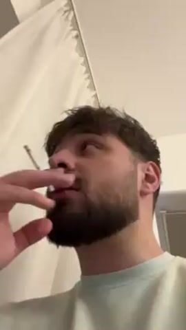 A man is holding a cigarette in his left hand and smoking it while looking down at the camera. He has short, dark hair and a full beard. He is wearing a light blue t-shirt. Behind him are white curtains and a white wall.