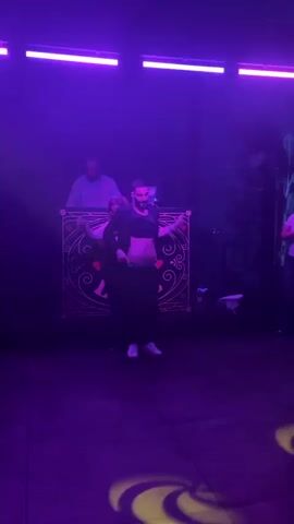 The video shows a man on a small dance floor, in front of a DJ booth, briefly exposing his abdomen to a woman who is standing behind him. The dance floor appears to be in a nightclub or similar venue. The man is wearing a suit jacket, a t-shirt, baggy pants, and white sneakers. The woman is wearing a dark-colored outfit with a blazer and glasses. In the background, a DJ in a striped shirt stands behind the DJ booth, which is decorated with a black and white design featuring hearts. The lighting in the venue is dark, with changing colors of red and purple creating a dynamic atmosphere. The man lifts up his t-shirt and jacket, briefly showing his midsection to the woman, then adjusts his clothes.