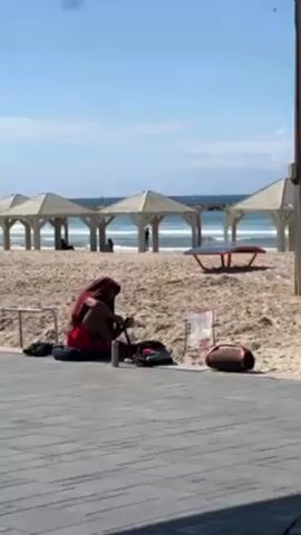 The video shows a person sitting on a tiled walkway next to a beach. They are wearing red shorts and what appears to be a head covering or hood. They seem to be focused on something in their lap, possibly manipulating or working with it. There is a backpack, a can, and a bag next to them, as well as a football or similar shaped object. In the background, there is a sandy beach with several structures providing shade. People are sitting under those structures. The ocean is visible in the distance under a bright blue sky with some clouds.