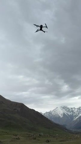 The video shows a black drone hovering in the air against a backdrop of cloudy skies and mountainous terrain. The drone is centrally positioned in the frame, and its propellers are spinning. The landscape includes a valley with green fields and snow-capped mountains in the distance. The foreground reveals a railing, suggesting the video was taken from an elevated location, possibly a balcony or terrace. The sky is overcast, creating a diffused lighting effect throughout the scene.