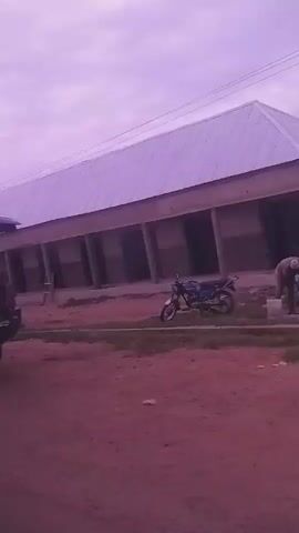 The video captures a side view of a dark blue vehicle, possibly a Volkswagen Golf, driving on a dirt road. The vehicle has a roof rack and appears to be older. In the background, there are buildings with corrugated metal roofs, a street light, and some vegetation. A motorcycle is parked in front of one of the buildings. The sky is overcast and has a purplish hue. The camera seems to be mounted inside another vehicle, as the view is shaky and the side of the interior of another vehicle is visible. The video appears to be shot during the day, but the lighting is dim. The environment seems to be a rural or semi-rural area. The video also includes brief glimpses of two other vehicles, one silver car and another blue car, as well as a person walking in the distance.