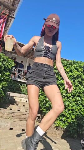 Two people, a woman and a man, are dancing outdoors on a wooden deck in what appears to be a party or event setting. The woman wears a brown Burberry cap, small sunglasses, a black lace-up top, black shorts, and black boots with white socks. She has tattoos on her arms and legs. The man is wearing a black shirt, pants with a white heart-shaped skull pattern, sunglasses, and a baseball cap. He's holding a clear plastic cup with a yellow liquid. A large green hedge forms the background, and a blue sky is overhead. There are other people visible in the background. The camera angle is low, looking up at the dancers.