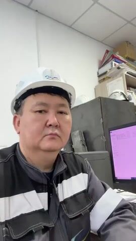 The video shows a man wearing a white hard hat and a grey work uniform with reflective stripes. He is sitting at a desk in what appears to be an office or control room. Behind him, there are shelves filled with files and boxes. To his right, a computer monitor displays a pink screen with text. The man is looking directly at the camera with a neutral expression. The lighting suggests an indoor environment with overhead fluorescent lights.
