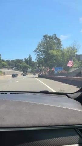 This is a first-person perspective video taken from inside a vehicle driving on a sunny day on a highway. The road ahead is clear with several cars visible in the distance. On the right side of the road, there is a sign that says "Tourist Information" with a blue directional arrow. American flags are also visible on the right side of the road, suggesting a patriotic or celebratory event. Trees and houses can be seen on the hills in the background. The dashboard of the car is visible in the foreground, with a textured surface. The sky is clear and blue.