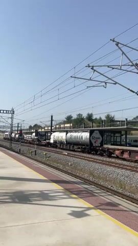 The video shows a train station platform with a train moving on the tracks. The train consists of multiple cars, including some tank cars with writing on them. The platform is paved with concrete and has a red and yellow striped border. There are overhead wires and poles supporting them, as well as traffic signals for the train. The sky is clear and blue, indicating a sunny day. Trees and buildings are visible in the background. The train appears to be traveling from left to right in the frame.