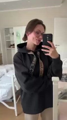 A young woman with brown hair pulled back in a low ponytail stands in a bedroom, holding a black iPhone in her right hand. She is wearing eyeglasses with rounded lenses and a black Calvin Klein Jeans hoodie with beige drawstring. She briefly sticks her tongue out. Behind her is a white bed with a partially made bed and a white dresser with mirrored doors. The walls are light beige.