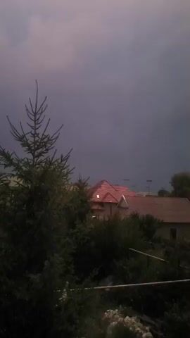 A silhouette of a coniferous tree is in the foreground on the left side of the frame. Behind the tree and to the right, several houses with pitched roofs are visible in the midground, with a single illuminated window in one of the houses. The background consists of a dark, overcast sky. The overall scene is dark and appears to be captured during twilight or at night.