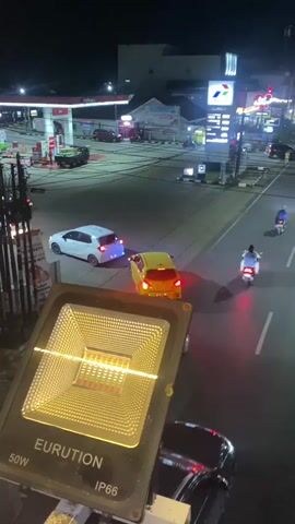 The video captures a nighttime street scene in Banjarbaru, Indonesia. The primary focus is a bright, square LED floodlight in the foreground, labeled "EURUTION 50W IP66." The floodlight emits a warm, yellow glow that illuminates the surrounding area.

Beyond the floodlight, a road is visible, bustling with activity. Two cars, one white and one yellow, are turning or maneuvering on the road. Red taillights from these vehicles and other vehicles further down the road indicate ongoing traffic. A motorcycle with a rider is also present, moving along the street.

In the background, a gas station with red and white pumps is illuminated by bright lights. Further in the distance, several buildings and signs are visible, including a prominent sign with a blue and white logo that appears to be for a petroleum company. The overall setting suggests an urban or commercial area at night. The lighting conditions indicate it is dark, and the scene is well-lit by artificial sources. There are no discernible emotions expressed by any visible subjects. The visible text includes brand names and specifications on the floodlight, as well as signs in the background which are not clear enough to read.