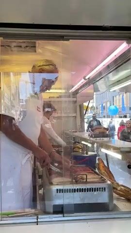 The video shows a food stand with a glass partition. Two people, dressed in white aprons, are working behind the counter. One person, appearing to be a man with blond hair, is facing away from the camera, and the other, appearing to be a woman with gray hair, is facing the camera and is tending to a grill with hot dogs. The grill is a silver, rectangular appliance with multiple slots for cooking sausages. There are several sausages on the grill, some of which appear to be hot dogs. In the background, outside the food stand, there is a crowd of people, some of whom are looking at the food stand. There are buildings and a blue balloon visible in the background. The overall setting appears to be an outdoor market or street fair.