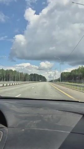 The video begins with a shot of a car's side mirror reflecting a red car driving on a road lined with trees and a guardrail. The camera then shifts to a close-up of a young woman with long blonde hair in a car. She has a red neck pillow and is wearing a white top. The woman blinks and sticks out her tongue at the camera before smiling.