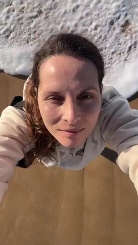 This is a first-person perspective video of a woman at the beach. The camera is pointed downwards towards the woman's face, with the ocean waves washing over her feet. The woman is wearing a light-colored hooded sweatshirt. The water is foamy and appears to be shallow, covering the sandy beach. The woman is looking directly at the camera and smiling. The video has a bright, natural lighting.