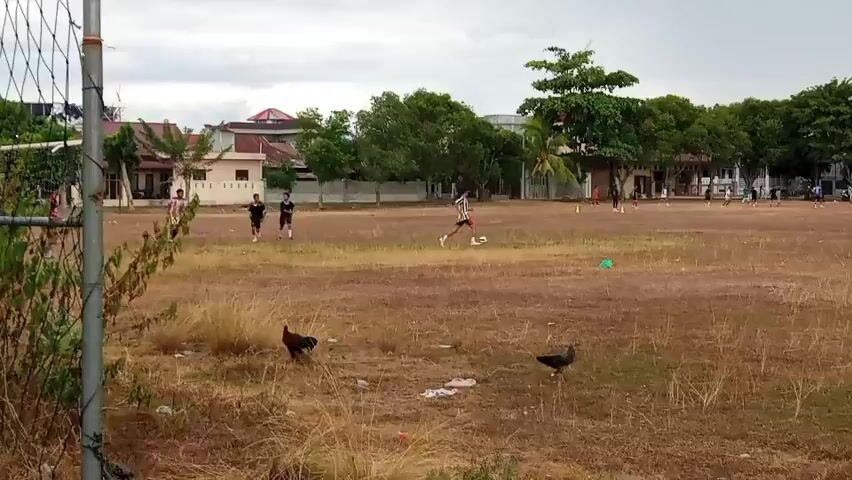 The video shows an outdoor scene on a dry, brown field with patches of yellow grass. In the foreground, two chickens are visible, one on the left and one on the right. Beyond them, a group of young people are playing soccer. One player in a black and white striped shirt and black shorts is kicking a soccer ball. Other players are running and interacting with each other on the field. In the background, there are buildings, trees, and a cloudy sky. There is also some debris scattered on the field. The overall environment appears to be a sports field or a large open area.