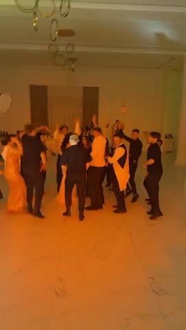 The video shows a group of people dancing in a brightly lit room. The room appears to be a venue with a large open space, possibly a ballroom or event hall. The floor is light-colored and reflective. In the background, there are draped curtains and some decorative elements hanging from the ceiling, including circular gold rings. The lighting in the room changes colors, cycling through pink, green, and red hues. Most of the people are dressed in semi-formal or formal attire, with many men wearing suits or dress shirts and trousers, and women in dresses or stylish outfits. Several individuals are holding their phones up, seemingly recording the event. The people are engaged in dancing, some in groups, others individually, with a general atmosphere of celebration and enjoyment. There are no obvious signs of distress or conflict.