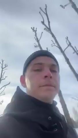 A person wearing a black beanie and jacket stands in a snowy, wintry environment. Behind them are several bare trees against a bright, overcast sky. The person is looking directly at the camera.