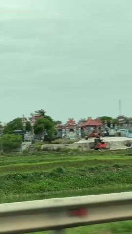 The video captures a scenic view of a cemetery with ornate structures and lush green vegetation. The sky is overcast, suggesting a cloudy day. In the foreground, a blurry white railing suggests the perspective of someone traveling in a vehicle, possibly a car or train. The cemetery features distinct architectural styles, with curved roofs and decorative elements on buildings that appear to be mausoleums or temples. There are also smaller grave markers scattered throughout the landscape. Adjacent to the cemetery, there's a patch of undeveloped land with some construction equipment, including an orange excavator, indicating ongoing work or development. The video pans across the scene, offering a continuous view of the cemetery and its surroundings.