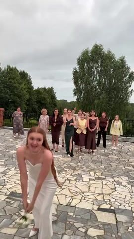 A bride in a white strapless dress throws a bouquet of white flowers backwards over her head towards a group of women standing in the background. The women are dressed in various outfits, mostly in shades of brown, green, and yellow. One woman in a dark green dress lunges forward and catches the bouquet as the bride turns and embraces her. The bride and the woman who caught the bouquet share a kiss. The other women in the background are clapping and cheering. The scene takes place outdoors on a stone patio with trees and greenery visible in the background under a cloudy sky. A person in the lower left corner is partially visible, appearing to be taking a photo with a camera.
