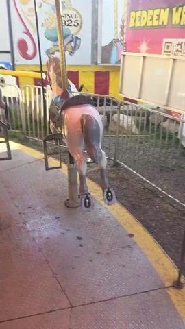 This video captures a carousel horse in motion at a fair or carnival. The horse is a light brown and white painted carousel horse, its hindquarters are most prominently featured. It is attached to a central pole and appears to be rotating. The carousel itself is adorned with a red and yellow canopy, suggesting an outdoor setting. 

The background reveals elements of a fairground or carnival environment. There's a fence enclosing the ride area, and behind it, the facade of a building with beige siding and yellow accents. A large mural-like painting on the side of a trailer or booth depicts a cartoonish character and the text "SINCE 1925," indicating the establishment's long history. Other carousel horses, partially visible, are also moving. In the distance, parked vehicles and trees are visible, and the bright sunlight suggests it is daytime. The overall impression is that of a lively and classic amusement park attraction.