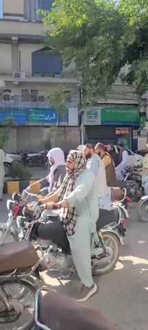 A group of men are riding motorcycles down a street in an urban setting. The men appear to be Middle Eastern or South Asian, many are wearing traditional clothing, including robes, turbans, or scarves. Some of the men are riding with children. The motorcycles are all older models. The street is lined with businesses, including banks. There are trees lining the street, providing shade. The weather appears to be sunny and warm. The video seems to be taken from street level and has the feeling of amateur footage.