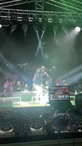 The video shows a live music performance. A bald, dark-skinned male performer is on stage, wearing a dark jacket, a light-colored t-shirt, and light-colored pants, holding a microphone and singing. He is standing in front of a large crowd of people, many of whom are looking at the stage. The stage is equipped with lighting and a DJ setup, including a laptop and DJ controllers. Smoke or fog is present on stage, illuminated by stage lights. The overall environment appears to be an outdoor or large indoor venue.