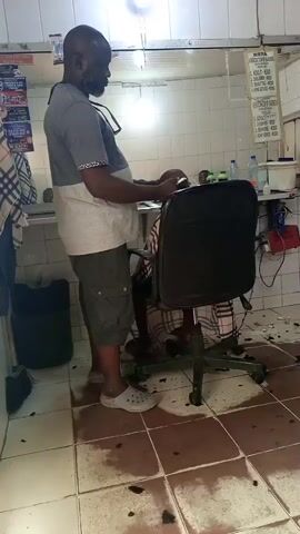 A barber is giving a haircut to a seated customer in a barbershop. The barber, a middle-aged Black man with a beard, is standing and wearing a two-toned t-shirt, cargo shorts, and white Crocs. His glasses are hanging on his shirt. The customer is seated in a black swivel chair and wearing plaid pants. Hair clippings are scattered on the tiled floor. The barbershop is small and has a mirror, a counter with various barbering supplies, and a sign with prices.