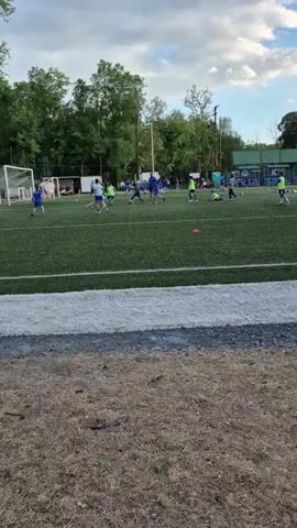 The video shows a soccer field with green turf, marked by white lines. Multiple players are spread across the field, most wearing blue jerseys, while a few are in neon yellow vests. There is a man in a black shirt standing on the field, near one of the goals, appearing to be coaching or observing. The players are actively engaged, moving around and interacting with each other. Orange cones are scattered across the field as part of a drill or practice setup. The background includes trees and greenery, with a hint of buildings or structures in the distance. The sky is overcast with scattered clouds. The camera remains static throughout the video, capturing the scene from a distance beyond a strip of dry grass and a painted white boundary line.