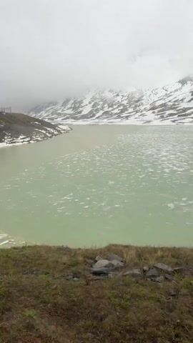 The camera looks through a train window out at a landscape. The foreground is a grassy ledge with a few visible rocks, beyond which is a pool of murky, greenish water. The water appears to have some thin ice or snow on the surface. In the background, a hillside is covered in snow and brown earth. Power lines and some small structures are visible on the hillside. The sky is gray and overcast. As the video progresses, condensation appears on the train window, and the scene becomes obscured.
