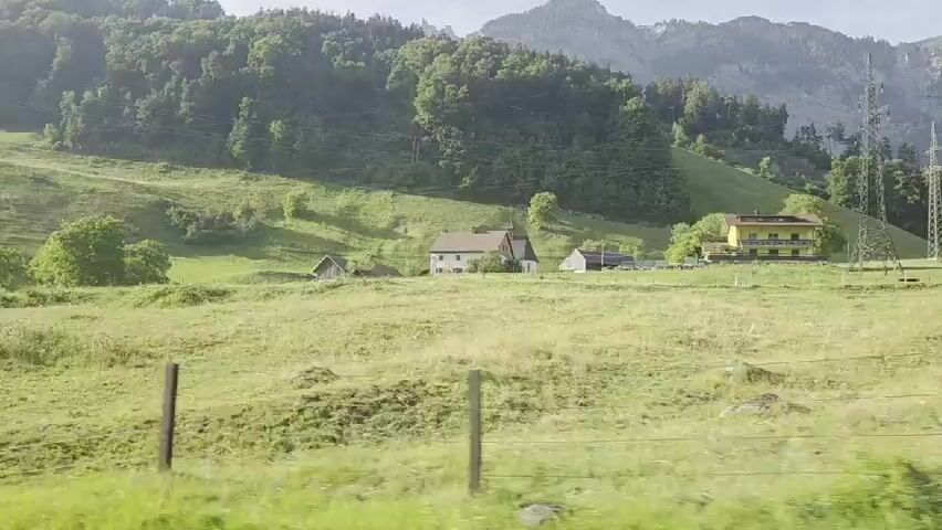 The video shows a rural landscape with rolling green hills and scattered houses. The foreground is dominated by a grassy field with wooden fence posts. In the middle ground, several buildings, including a white house with a red roof and a yellow building, are visible. The background features a densely wooded hillside leading up to a majestic mountain range. A tall electrical tower is also present on the right side of the frame. The sky is bright and appears to be daytime. The camera appears to be in motion, likely from a vehicle, as indicated by the slight blur and consistent movement across the scene.