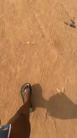 The video shows a dirt road leading toward a two-story building with a brown roof. The building is painted with peach and off-white colors. There is a water tower behind it. A black car is parked on the side of the road. The road is dusty and has tire tracks. The sky is overcast.