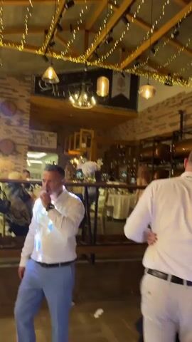 The video appears to be shot at a wedding reception. People are dancing on the dance floor, illuminated by blue stage lights, and other guests are standing around tables. The venue has exposed brick walls and string lights hanging from the ceiling. There’s a mix of adults and children present, some dressed in formal attire. The atmosphere seems festive and lively.