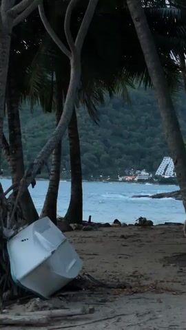 This media file captures a serene beach scene in Rawai, Thailand, likely during the late afternoon or early evening. The foreground is dominated by the sandy shore, with debris and fallen leaves scattered across the ground. A small, white boat is overturned on the sand, leaning against the base of a large palm tree. The texture of the sand and the weathered appearance of the boat suggest a sense of stillness and perhaps abandonment.

Behind the beach, lush green palm trees frame the view, their fronds creating a dappled canopy. The ocean stretches out in the middle ground, its waters a tranquil blue-green, with gentle waves lapping at the shore. In the distance, a verdant, tree-covered hillside rises, creating a dramatic backdrop. Nestled on the hillside is a distinctive, stepped white building, possibly a resort or hotel, its geometric form contrasting with the natural landscape. The building is illuminated, indicating that dusk is approaching or has already begun.

The overall atmosphere is peaceful and tranquil, with a hint of melancholy conveyed by the overturned boat. The lighting suggests a soft, diffused light, typical of twilight. No people are visible in the frame, contributing to the sense of quietude. The location cues—palm trees, lush vegetation, the distinctive architecture in the background, and the mention of Rawai, Thailand—clearly establish the setting. There is no visible text in the media file.