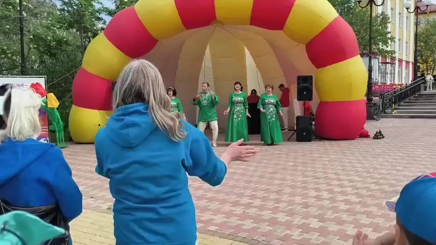 A group of five people, four women and one man, are performing in front of an inflatable archway. The women are wearing long green dresses with intricate embroidery, while the man is dressed in a white shirt with a similar design and loose-fitting pants. Two of the women and the man appear to be singing or speaking, with the other two women standing beside them. The archway is red and yellow, and it is positioned in an outdoor paved area. In the foreground, a woman in a blue hoodie and sunglasses is dancing, facing away from the camera. To her left, another person is partially visible, wearing a blue jacket. To the right of the stage, there are speakers and a red structure. In the background, trees and buildings are visible. A sign with red tulips is also present on the left side of the frame. The ground is paved with interlocking stones in a patterned arrangement. The overall scene suggests an outdoor performance or festival.