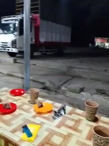 A dimly lit outdoor scene at night. In the foreground, a table is covered with a patterned tablecloth. On the table are two red plates, one orange plate, a pack of cigarettes, two cups, a lighter, and some food items. In the background, two white trucks are parked. One truck is closer to the foreground and is facing the camera. The other truck is further back and is partially visible. There is a pole in the middle of the scene. The ground is paved with concrete.