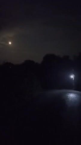A handheld, shaky, low-light video captures a dimly lit, rural road at night. The road appears to be paved and wet, reflecting the light from a single, bright lamp post in the distance. The sky is dark with clouds, and a full moon is visible in the upper left corner, partially obscured by clouds. The surrounding landscape is dark, with silhouetted trees and vegetation lining the sides of the road. The camera is moving forward, creating a sense of traveling down the road. There are no people or animals visible in the video. The overall atmosphere is quiet and slightly eerie.
