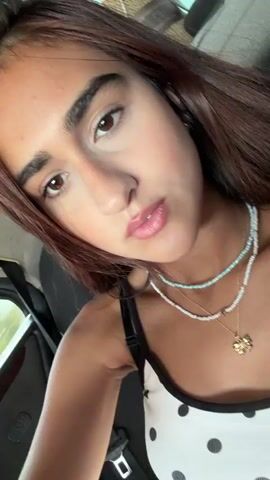 The video is a close-up selfie of a young woman with long, dark hair and light skin. She is wearing a white tank top with black polka dots and two necklaces, one with light blue and white beads and another with a gold pendant shaped like a four-leaf clover. She is also wearing a black bra underneath her tank top. The background is blurry but appears to be the interior of a car, with a seatbelt visible on the right side of the frame. The woman is looking at the camera with a neutral expression, and her lips are slightly parted. The lighting is soft and natural.
