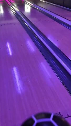 The video begins with a close-up of a bowling ball with a hexagonal pattern, resting on a bowling lane. The lane is illuminated with purple lighting. The camera then pans up the lane to show pins at the far end. The ball rolls down the lane towards the pins. The camera then cuts to a wider shot showing multiple bowling lanes with neon numbers above them, indicating lane numbers 11, 12, and 13. A bowling ball is seen on lane 11, heading towards the pins. The video concludes as the ball strikes the pins.