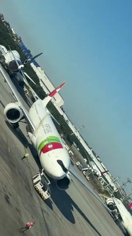 A white airplane with a green and red stripe on its body and tail is parked on an asphalt tarmac on a sunny day. A yellow tug vehicle is attached to the nose of the plane, and two ground crew members in yellow vests are standing near the plane. In the background, other airplanes are visible, along with buildings and trees. The sky is clear and blue.
