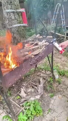 A person is using a canister with a red nozzle to spray something onto a fire in a metal barbecue grill. The fire is burning brightly with flames reaching high. The grill is set up outdoors, near a tree and some greenery. There are logs and branches in the grill, some of which are already burning. In the background, there's a ladder, a fence, and more trees, suggesting a rural or suburban setting. The ground is covered with dirt, leaves, and some paving stones. A plastic bottle is visible on the ground near the grill. The lighting suggests it could be daytime, possibly overcast or late afternoon. The overall scene depicts someone preparing to barbecue or rekindle a fire.