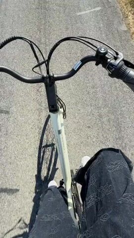 A first-person perspective video shows someone riding a bicycle on a paved road through a rural landscape. The road is lined with green grass and trees. The sky is clear and blue. In the distance, there are houses and hills. The video appears to be taken on a sunny day.