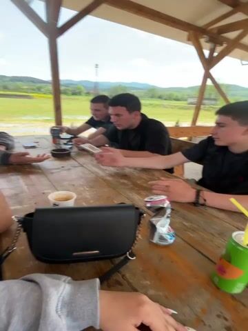 The video shows a group of young men seated at a wooden picnic table under a covered structure, seemingly engaged in a card game. One of them smokes a cigarette while another drinks from a can. A cup of coffee sits on the table, alongside other personal items like a black handbag and another crushed can. The outdoor setting includes a grassy field, distant hills, and a blue sky with scattered clouds. 

Subsequently, a person walks a small dog on a leash outside the covered area. Two young women then appear, posing for the camera with pouty faces under the same covered structure. The final scene shows the same small dog, still on its leash, sniffing the ground outside.
