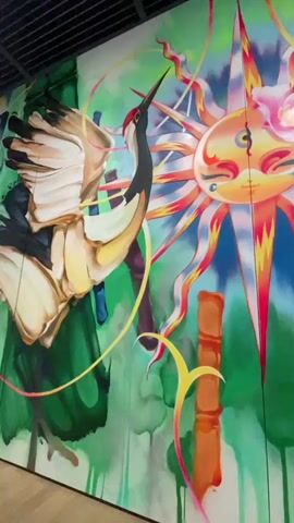 The video displays a mural with a vibrant, psychedelic aesthetic. A crane with outstretched wings is depicted on the left, its feathers detailed with shades of white, brown, and black. To the right of the crane is a stylized sun with a human-like face, complete with closed eyes, blushing cheeks, and a pink flower adorning its head. The sun's rays are rendered in a spectrum of colors, radiating outwards. In the background, abstract green forms resembling bamboo stalks and flowing water are visible. Various elements, including a coiled snake and a leopard or snow leopard, are interspersed throughout the mural. The overall impression is one of a dreamlike, possibly spiritual, landscape.