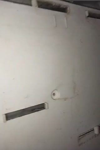 A hand is visible in the foreground of a close-up, shaky, blurry video of the interior of a refrigerator or freezer. The hand is reaching into the compartment where a coil system, wires, and what appears to be a black component are located. The compartment is white and shows some signs of dirt and possible water residue along the bottom edges. The coil system is metallic and appears to be a heat exchanger. The video pans slightly, revealing more of the interior walls of the appliance.
