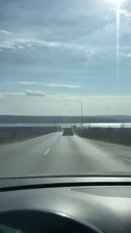 A first-person perspective video shows someone driving on a highway. The road is straight and has other cars ahead. There is a body of water on either side of the highway in the distance, illuminated by the sun. The sun is high in the sky, creating a lens flare effect. The dashboard and steering wheel of the car are visible. The driver's hand is visible holding the steering wheel at times. There are streetlights along the side of the road. A phone mount is visible on the dashboard.